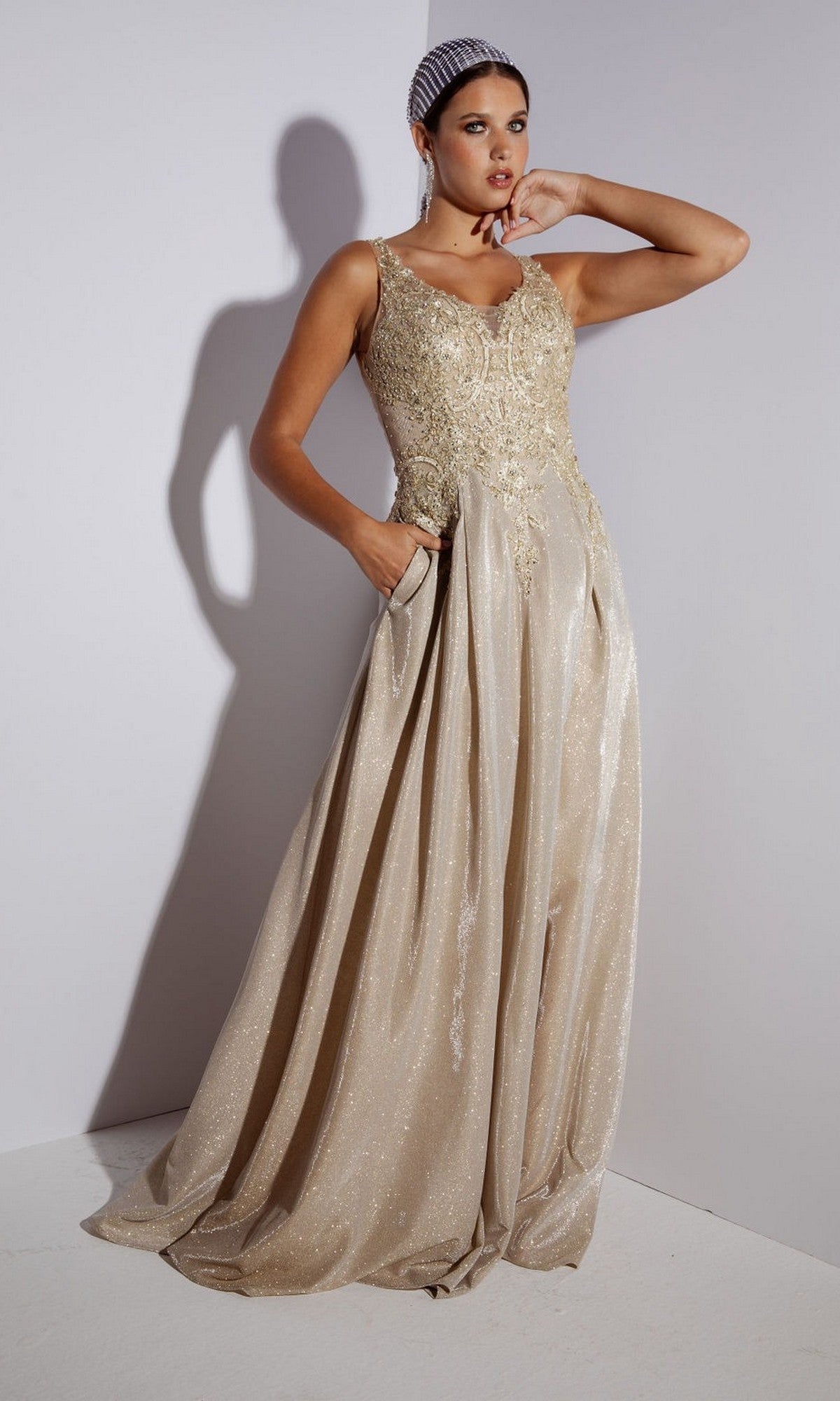 A woman in the Eureka 9606 Long Prom Dress, featuring a mocha color, sleeveless design, embroidered bodice, and shimmering A-line skirt, poses gracefully against a light-colored wall.